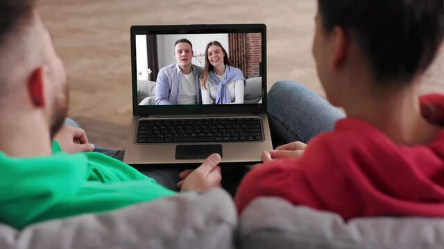 Zoom out. Caucasian Couple Using video call at home to connect with friends at home. Shot behind models shoulders. Back view of young atractive couple boy and girl sitting at home and speaking online.