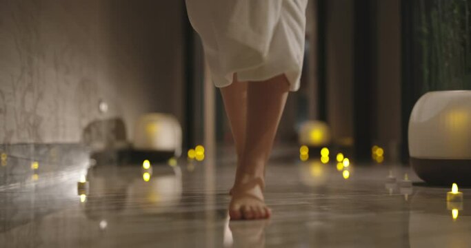 Close up shot of slender female legs slowly stepping on floor with candles. Woman in white robe walking in resort spa low angle 4k footage