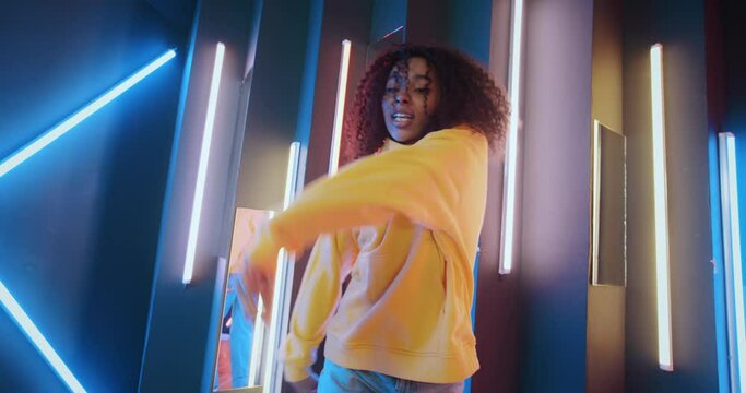 Handheld wide angle shot beautiful black woman dancing modern dance on colorful striped lights background. Attractive African American urban female wearing yellow hoodie in night disco club