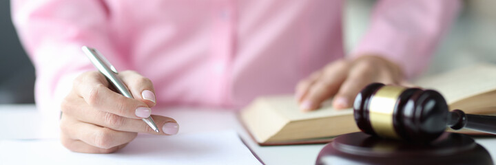 Woman judge writing in documents with ballpoint pen near hammer of courthouse