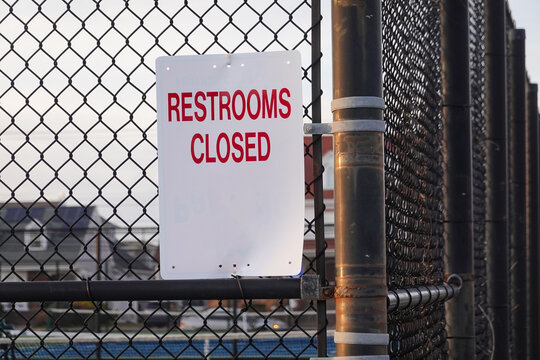 White sign with red lettering that says, Restrooms Closed on a metal chain link fence by a playground
