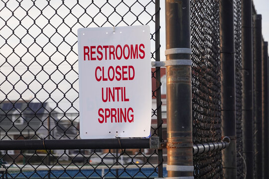 White sign with red lettering that says, Restrooms Closed Until Spring on a metal chain link fence by a playground