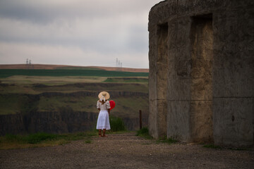 woman in white dress with straw hat facing away standing alone with a red balloon at the stonehenge...