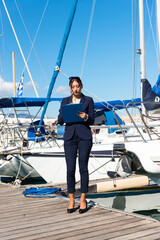 Female yachting dealer in marine blue suit holding notepad, standing outside a luxury sailboat,...