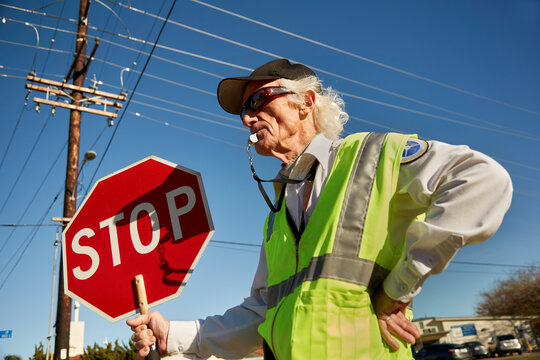 A child traffic controller stands in the street with a stop sign, capy, sunglasses and yellow vest. Low shot. Man with gray hair looks to the left his head shadow is on the stop sign. Day.background p