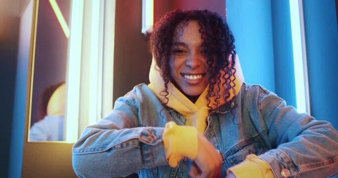 Cheerful black woman dancing modern dance on colorful striped lights background. Urban female wearing yellow hoodie jeans jacket sitting on floor moves rhythmically to music beat in night disco club