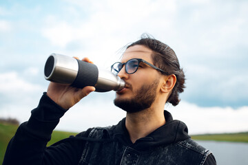 Portrait of young man drinking water from reusable steel thermo bottle on background of blurred cloudy sky. Wearing glasses. - obrazy, fototapety, plakaty