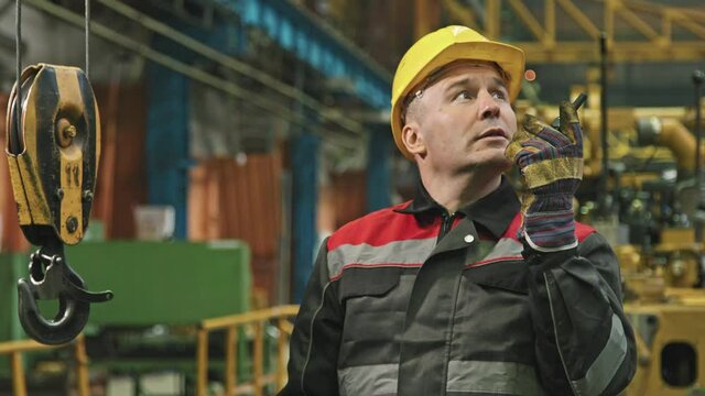 Medium shot of adult male plant foreman in yellow hard hat talking on walkie-talkie controlling tractor assembling process at factory