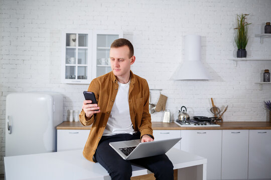 An attractive young man in casual clothes sitting in the kitchen using a laptop computer. Work from home, remote workflow.