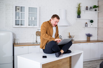 An attractive young man in casual clothes sitting in the kitchen using a laptop computer. Work from home, remote workflow.