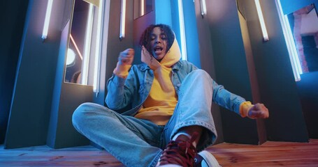 Stylish urban female wearing yellow hoodie and jeans jacket sitting on floor moves rhythmically to music beat in night disco club. Handsome black woman dancing on colorful striped lights background