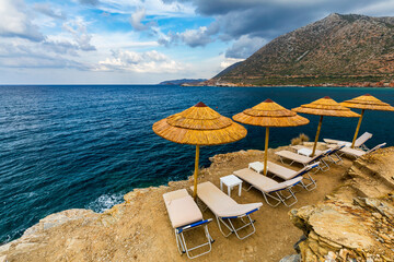 Summer vacation destination. Straw sunshades and sunbeds on the empty rocky beach with sea in the background. Vacation And Tourism Concept. Sunbeds On The Paradise Beach. Umbrellas and sunbeds.
