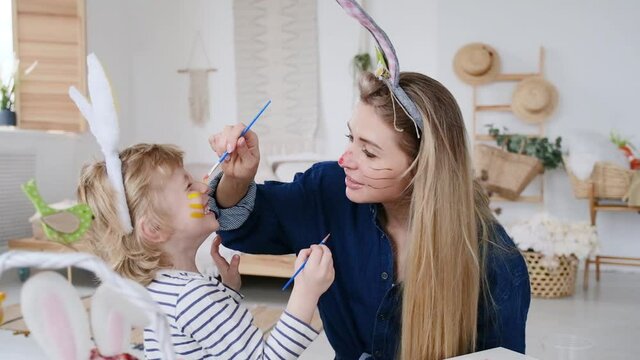 Beautiful happy son and mother in headbands with bunny ears decorating eggs using paintbrush and bright colors of paint, preparing to celebrate Easter holiday, having fun spending time together at hom