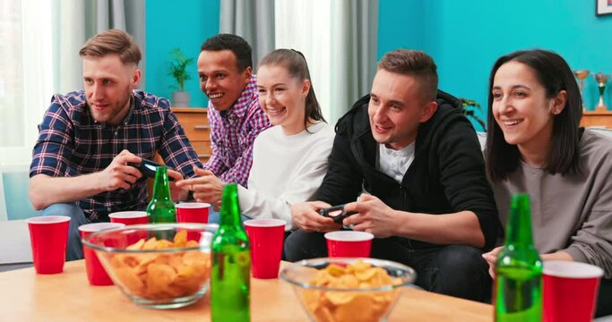 A multinational group of college students sit on the couch and play video games on the TV at home. Smiling friends hang out. The boys compete in a video game. One of the players is cheating