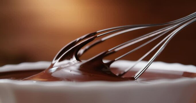 Close-up chocolatier mixing melted liquid chocolate with steel whisk. Hot chocolate mix in white bowl on wooden background. Cooking handmade chocolate dessert and candies. Confectionery concept