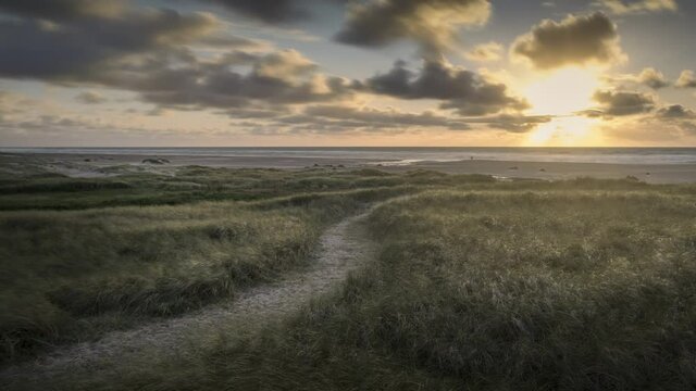 Time lapse of pathway in sand dunes during sunset on coast of Denmark, clouds moving in sky, people walking on beach
