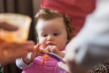 Portrait of a little blond toddler girl. Spoon eating. Dirty mouth. Mom feeds the baby.