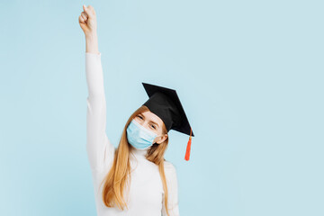 Excited female graduate in graduation hat and medical mask celebrating success against blue background