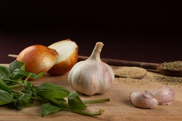 Wooden cutting board with vegetables or cooking ingredients; whole head of garlic and cloves, one onion in half, basil leaves and two wooden spoons with pepper and oregano; dark background.