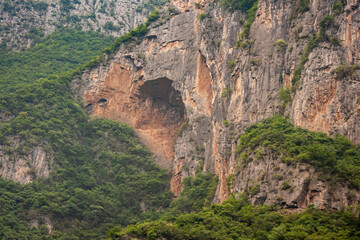 Fototapeta na wymiar Guandukou, Hubei, China - May 7, 2010: Wu Gorge on Yangtze River. Closeup of Giant cave in red-gray rock cliff of forested mountain.