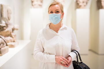 Adult woman visitor in protective mask looking at exposition of artworks in historical museum