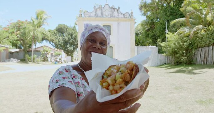 Happy Brazilian woman dressed in the traditional Bahian costume of the Umbanda religion, offering acaraj&eacute;, typical Bahia food in the historic center of Porto Seguro, Brazil. 4K.