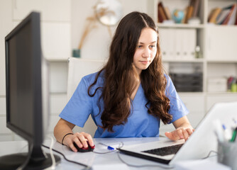 Young female trainee doctor who is undergoing an internship at a medical clinic is sitting at workplace at a computer