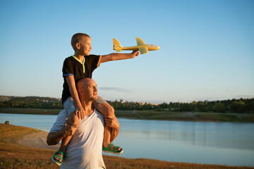 Father holds his son on his shoulders against the backdrop of nature by the lake at sunset. The boy is holding the plane in his hand. Family vacation concept