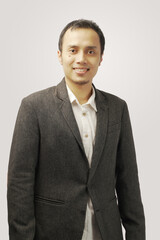 Smiling young man in dark suit crossing hands on chest and looking at camera while standing against gray background. 