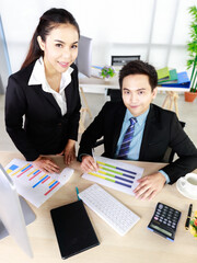 Asian man sitting holding hand pen with A woman standing in a black suit in the office on the table there are pencils, flowers, note books and computers. Concept business for working.