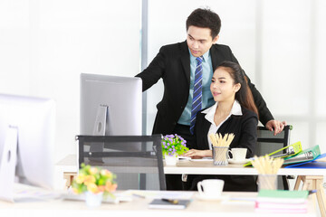 Asian businessman standing guides working with smiling businesswoman using a desktop computer and business graph file document is placed on their desk in the office. Concept team meeting for work.