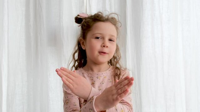 Funny little girl in pink clothes shakes head with space buns and shows stop gesture standing near white curtains in light room