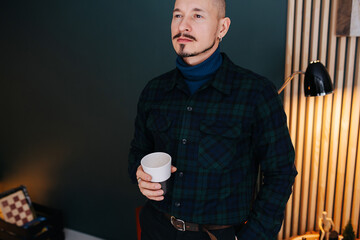 Proud serious middle aged man holding a mug, having a warm drink at home.