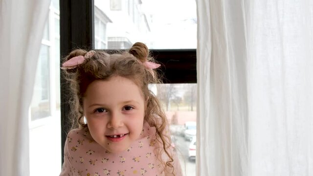 Adorable little girl with small space buns on head and lost tooth talks to camera playing with curtains in light room closeup