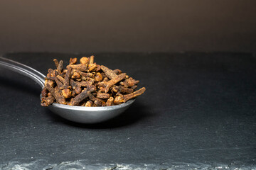 metal teaspoon full of cloves, isolated on dark background, black stone texture, macro shot