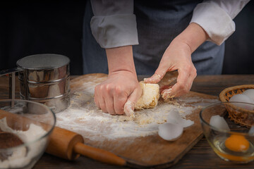 Women's hands, flour and dough. A woman in an apron cooking dough for homemade baking, a rustic home cozy atmosphere, a dark background with unusual lighting.