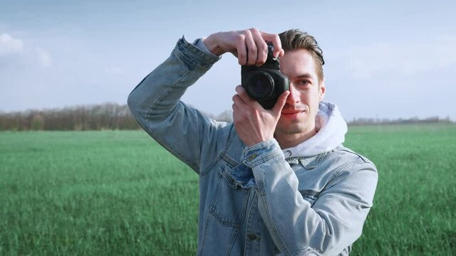 Young male photographer taking photo on professional photocamera, standing at beautiful green field. Portrait of man using digital camera outdoors