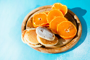 On a round wooden cutting board, there are round pancakes and round orange slices. Blue background. Copy space for text.