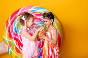 Obraz na płótnie Canvas two happy little girls in colorful dress drinking orange juice having fun on yellow background with lollipop