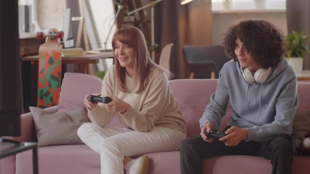 Beautiful woman and her teenage son smiling and using controllers while playing console game on sofa at home