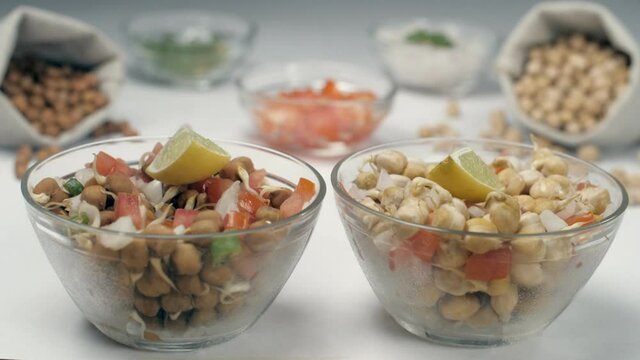 Two lemon slices falling inside a bowl of  sprouted chickpeas salad mixed with onions . Two bowls of chopped tomatoes and green chilies kept together against a blurred background