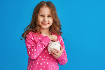 Fototapeta na wymiar Happy savings. A little cute girl with a piggy bank on a blue background. The child smiles happily and puts money in the piggy bank The concept of saving money for a dream.