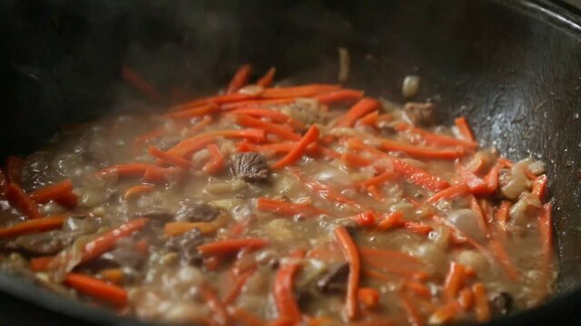 Meat, carrot and onions are stewed in a cauldron in a sauce close-up. The cook pours spices for pilaf, cumin, barberry, salt. Cooking pilaf in a cast-iron kazan, zirvak, oriental cuisine. Step 6 of 8
