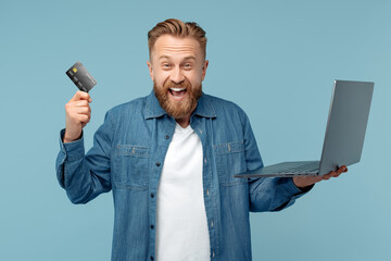 Cheerful bearded hipster blonde young man in jeans shirt holding notebook and credit card over blue...