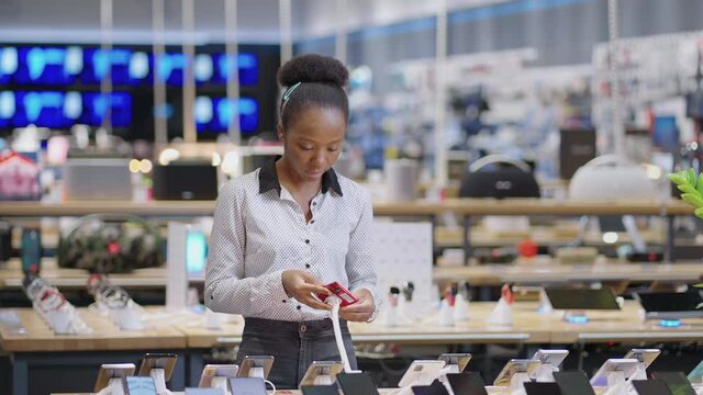 black female shopper is viewing exhibition samples of modern mobile phone in electronics shop, lady is buying smartphone in retail shop