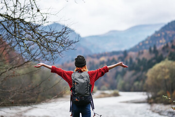 a traveler in a hat, sweater and trousers gestures with her hands on the river bank in the mountains