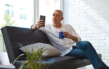 Satisfied man relaxing in his living room using mobile phone while having coffee