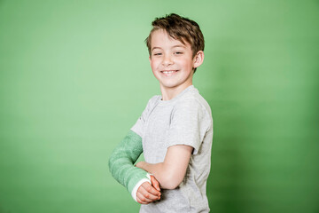 cool young schoolboy with broken arm and green plaster posing in front of green background in the studio