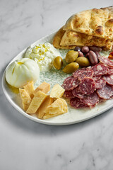 Italian antipasti snacks on white plate on marble background. Focaccia, burrata, olives, sun-dried tomatoes, salami, cheese, parmesan.