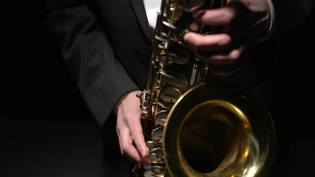 Musician in formal suit plays shiny saxophone with hands in the studio, close-up on a black background. Male saxophonist plays with fingers on the valves of a gold saxophone.